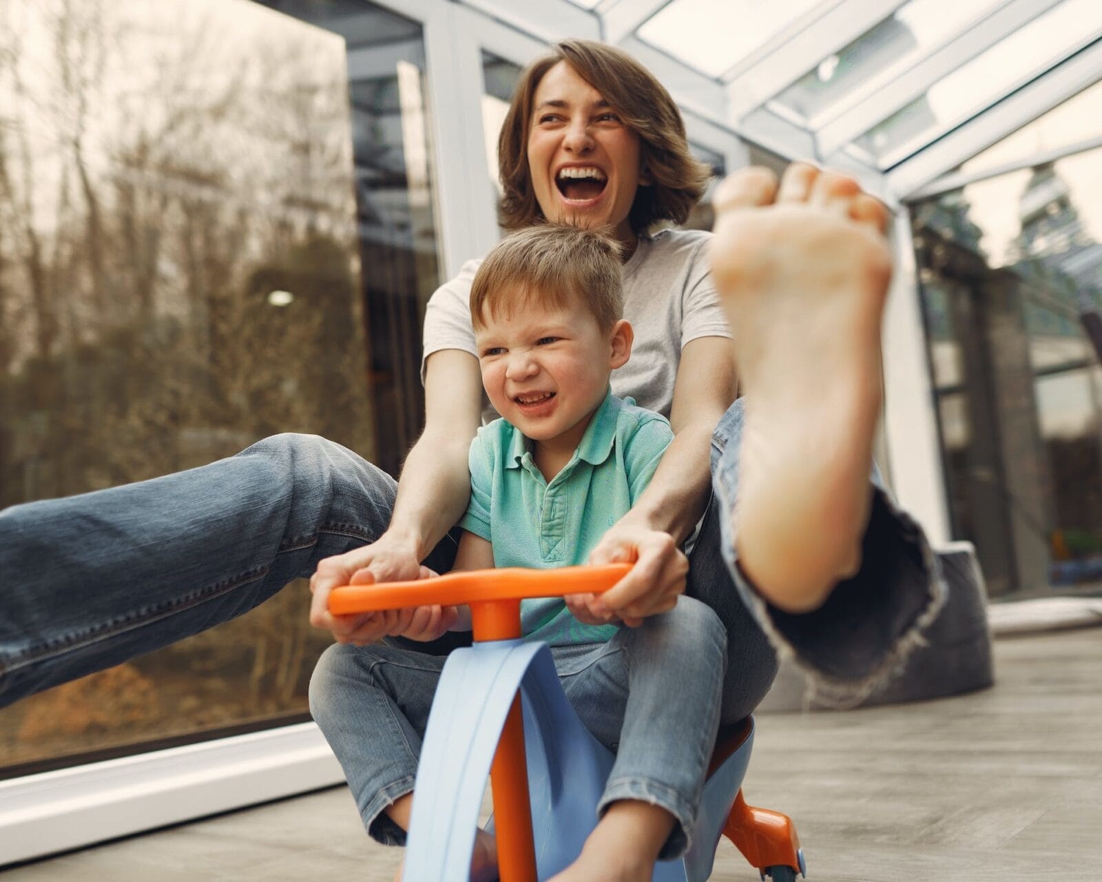 Laughing woman and young boy riding a toy tricycle indoors, enjoying playful and happy moments together.