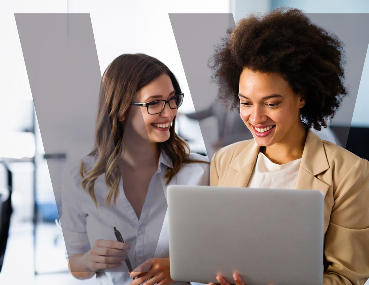 Two women smiling while working together on a laptop in a bright, modern office.