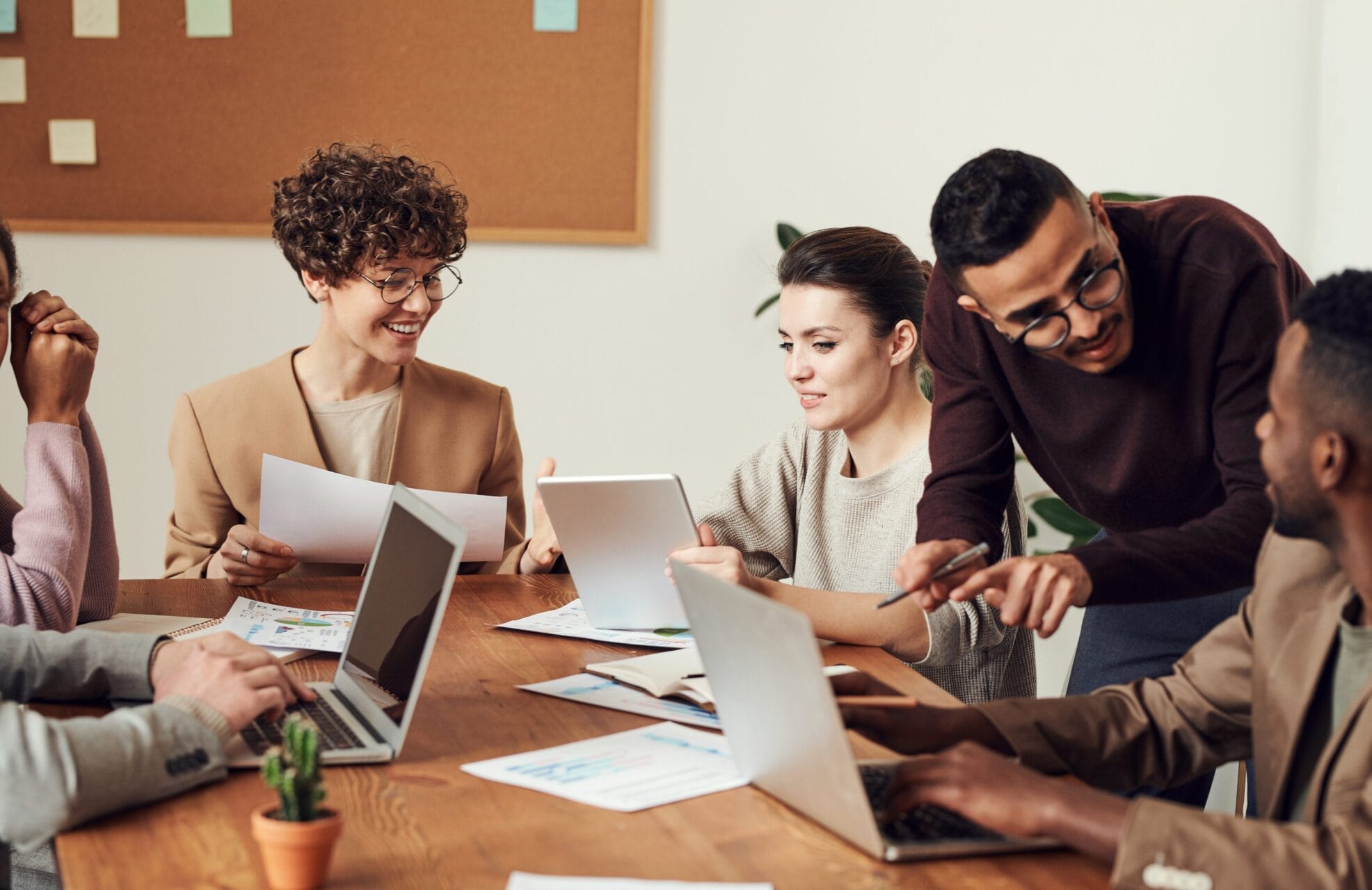 Group of diverse colleagues collaborating at a wooden table with laptops, documents, and tablets in a casual office setting.