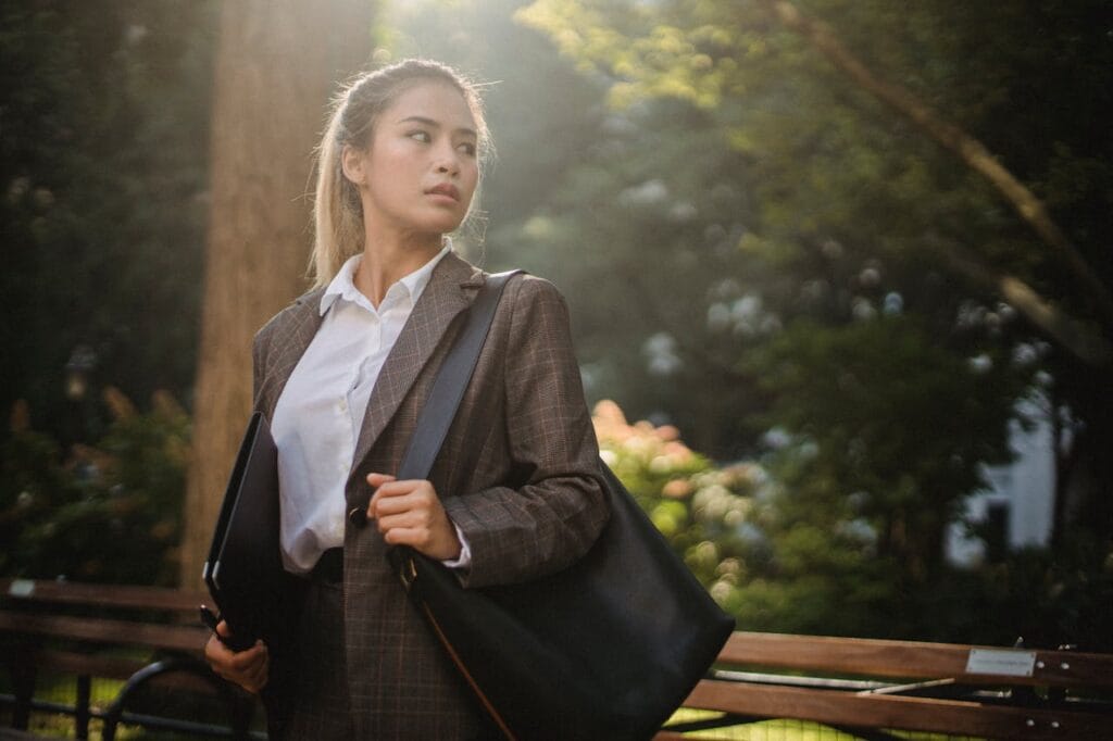Professional woman in a business suit standing outdoors in a park, holding a bag and looking to the side.