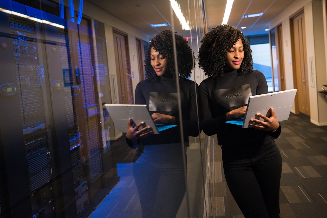 Woman standing in a server room, holding and working on a tablet, with her reflection visible on a glass panel.
