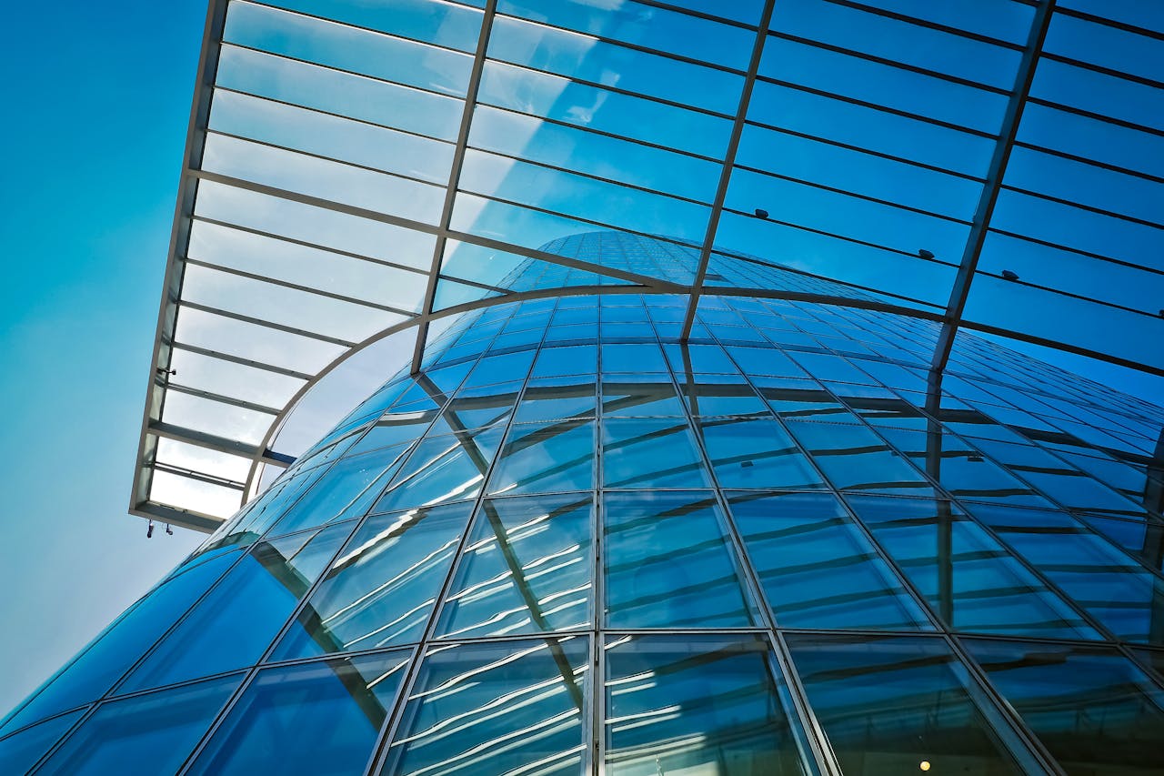 Low-angle view of a modern glass building with a curved facade and a metal framework under a bright blue sky.