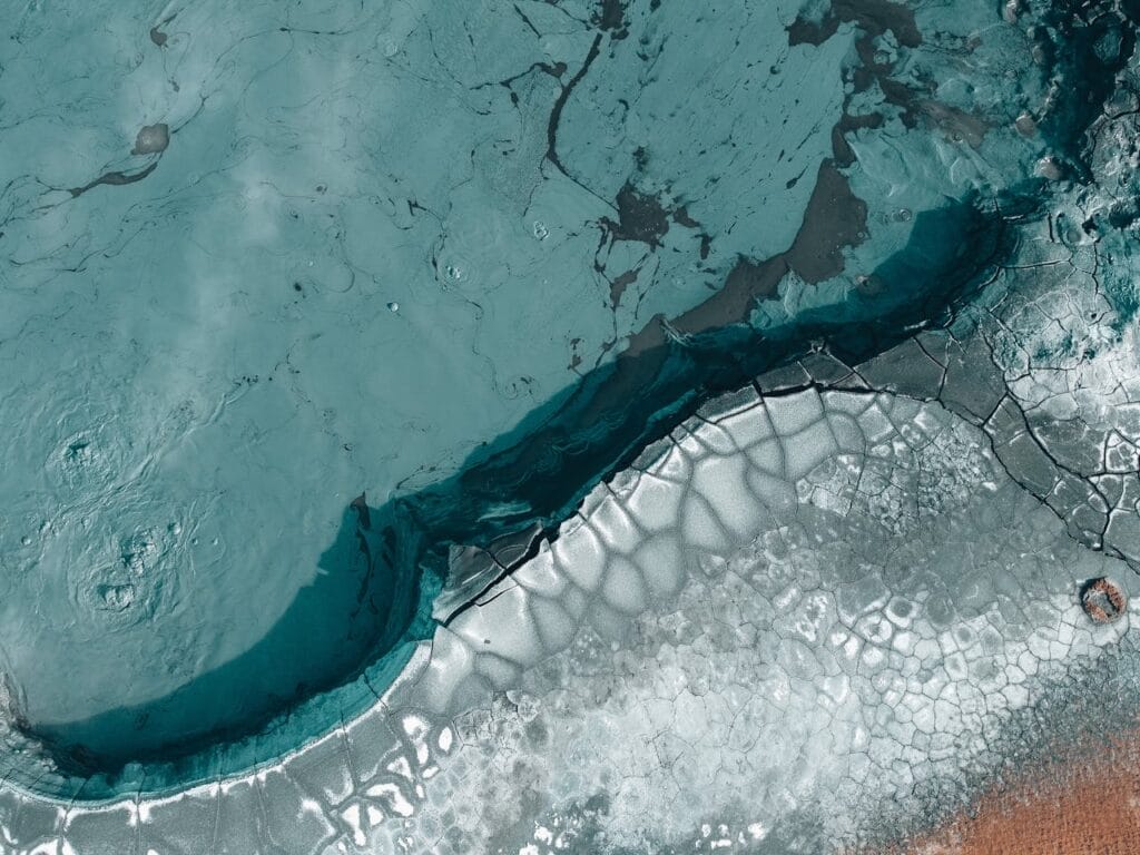 Aerial view of icy and cracked terrain meeting a body of turquoise water, showing natural patterns and textures.