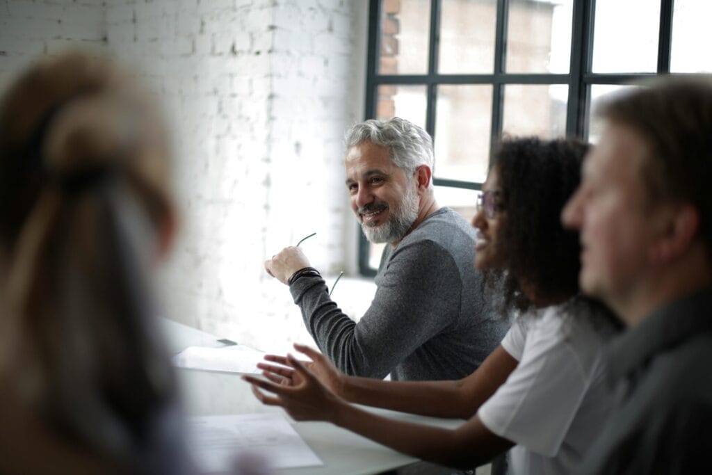 Group of diverse colleagues sitting at a table in a bright office, with one man smiling and holding glasses.