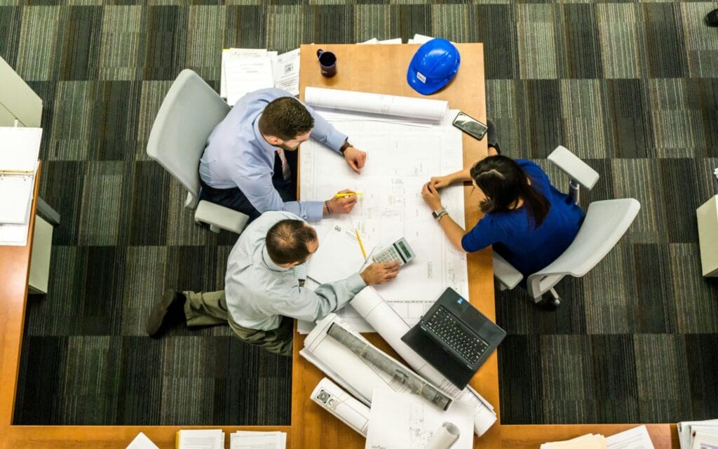 Top-down view of three people collaborating on a blueprint at a desk, with a laptop, calculator, and a blue hard hat on the table.