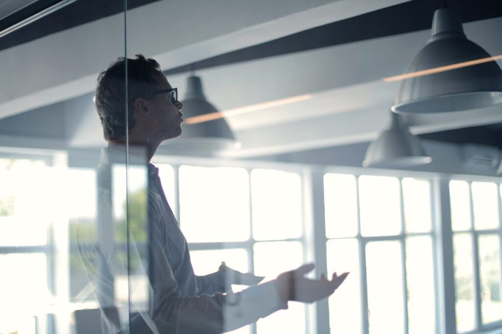 Man in a modern office standing behind a glass wall, gesturing with his hands during a presentation or discussion.